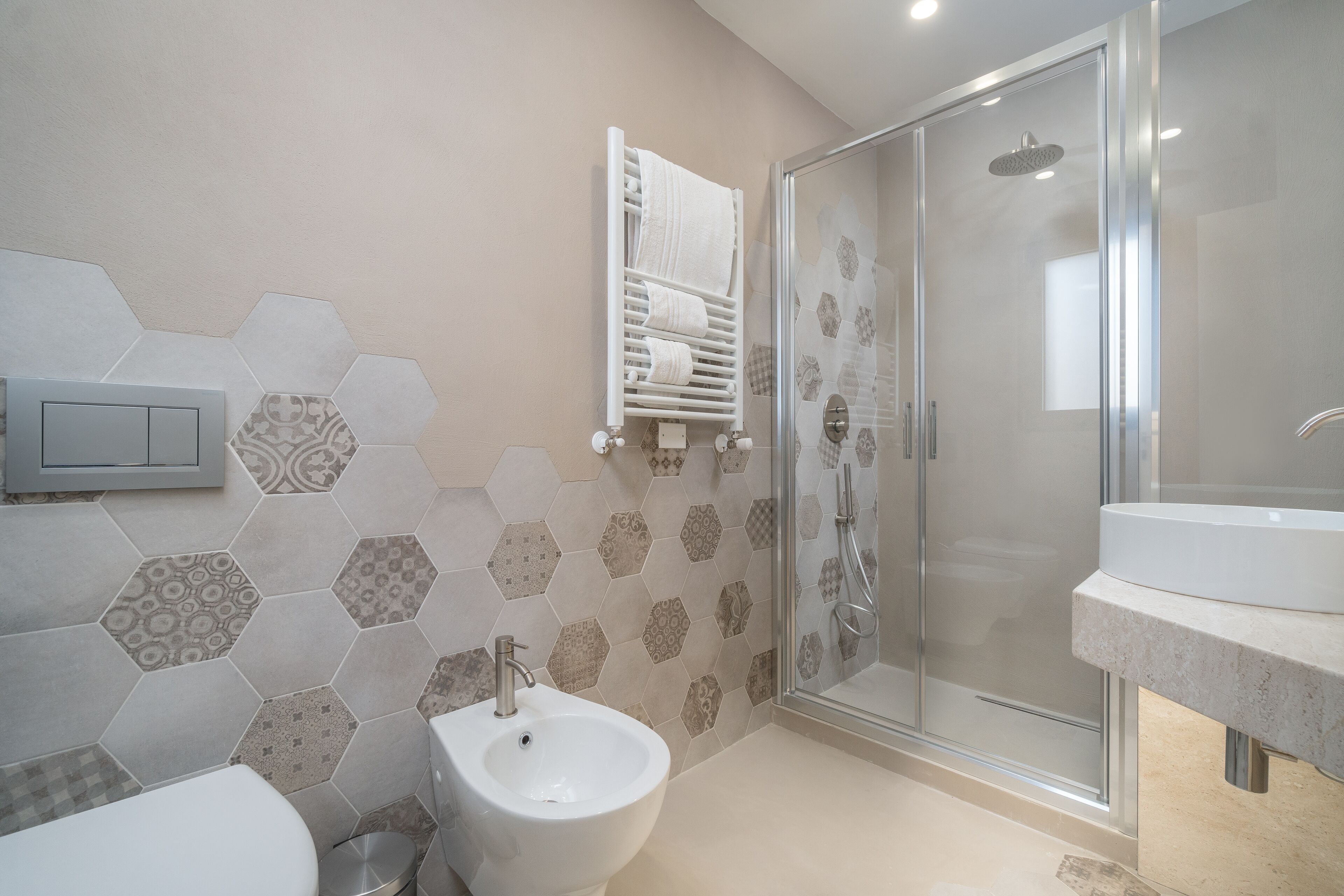 A modern bathroom with a hexagonal tile backsplash, a white sink, a bidet, and a glass-enclosed shower.
