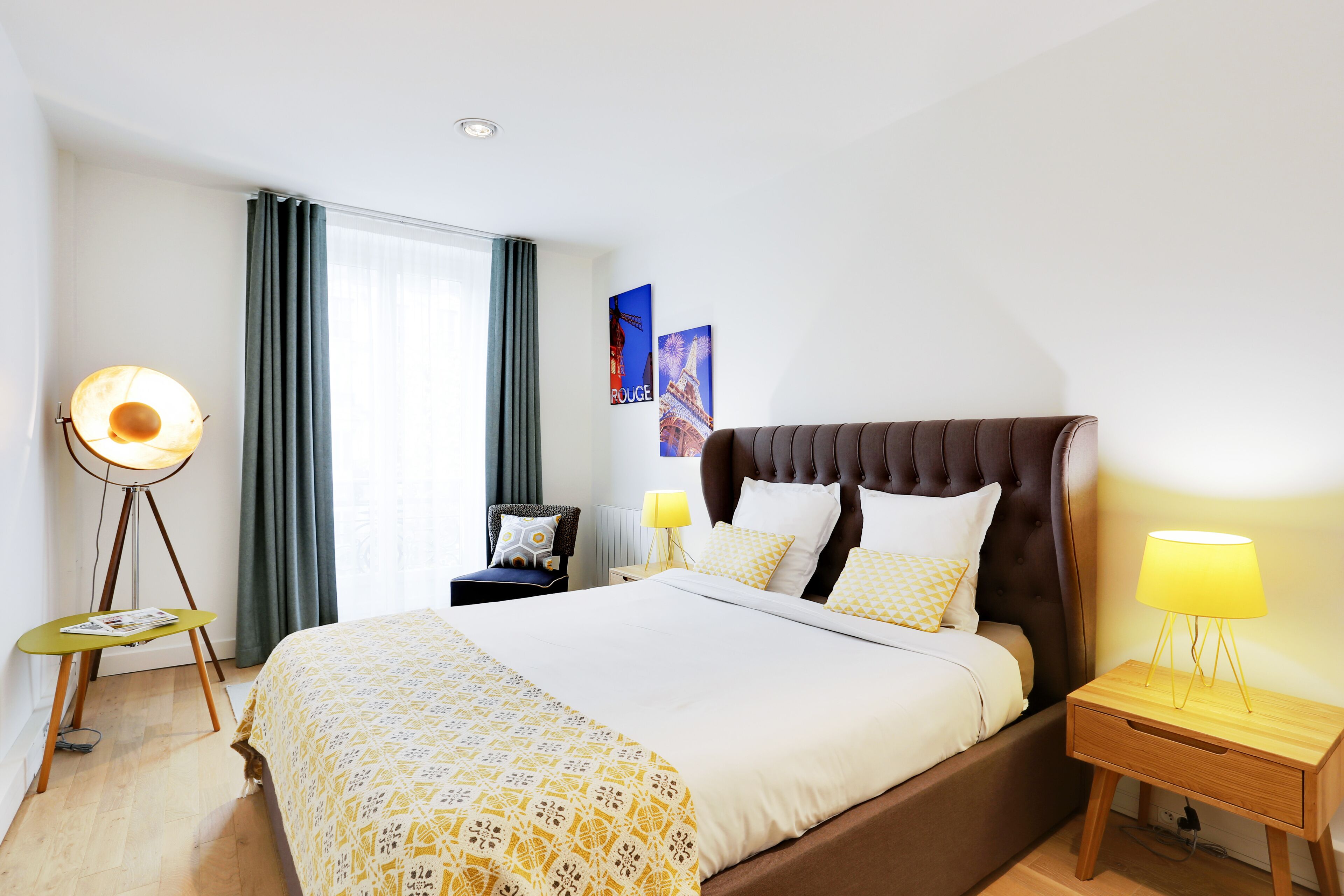 A modern hotel room with a large bed, a bedside table with a lamp, a floor lamp, and a view of the Eiffel Tower through the window.