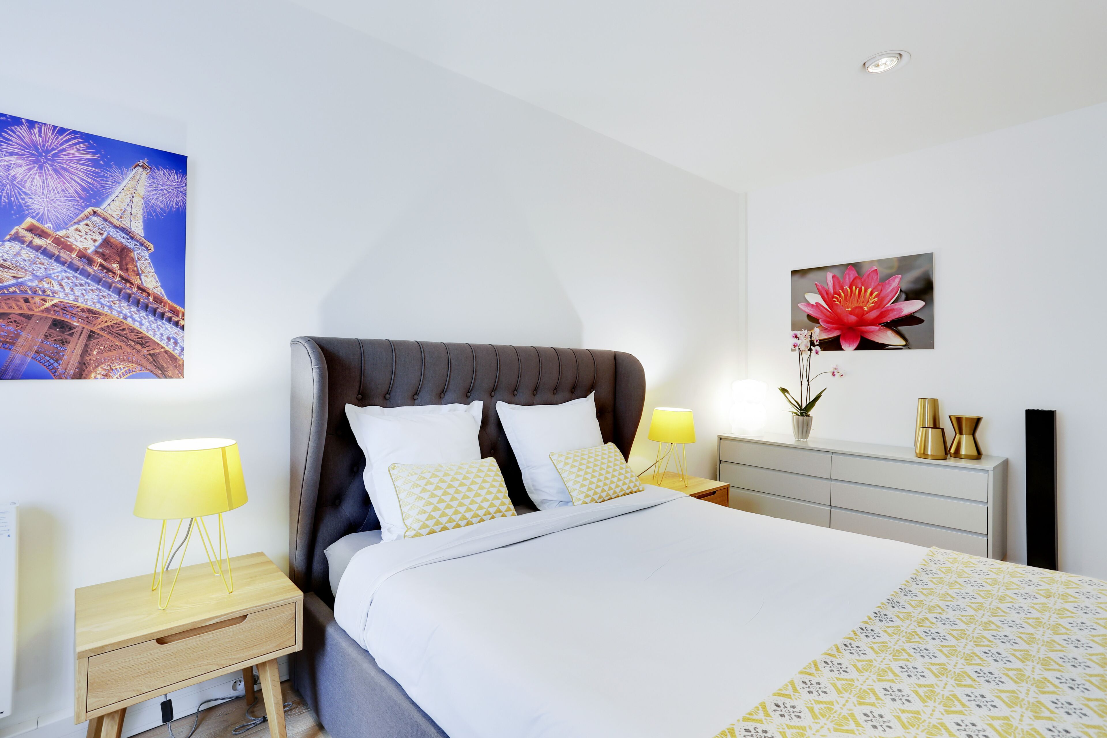 A bedroom with a bed, two bedside tables, a nightstand, a dresser, and a wall with a large picture of the Eiffel Tower and a flower.