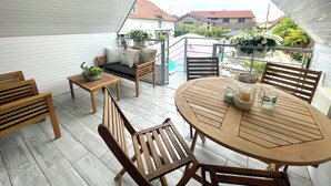 Une terrasse extérieure couverte, avec une table et des chaises en bois, un banc rembourré et une balustrade donnant sur un jardin.
