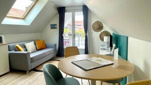 Une pièce chaleureuse de style loft, avec un plafond haut, une lucarne, un canapé gris agrémenté de coussins orange, une table ronde en bois et un fauteuil en osier.
