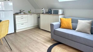 Une cuisine compacte avec des meubles blancs, un four à micro-ondes et une cuisinière à gaz. On y trouve un canapé gris avec des coussins jaunes et blancs, ainsi qu’une chaise en osier. Une lucarne apporte de la lumière naturelle.