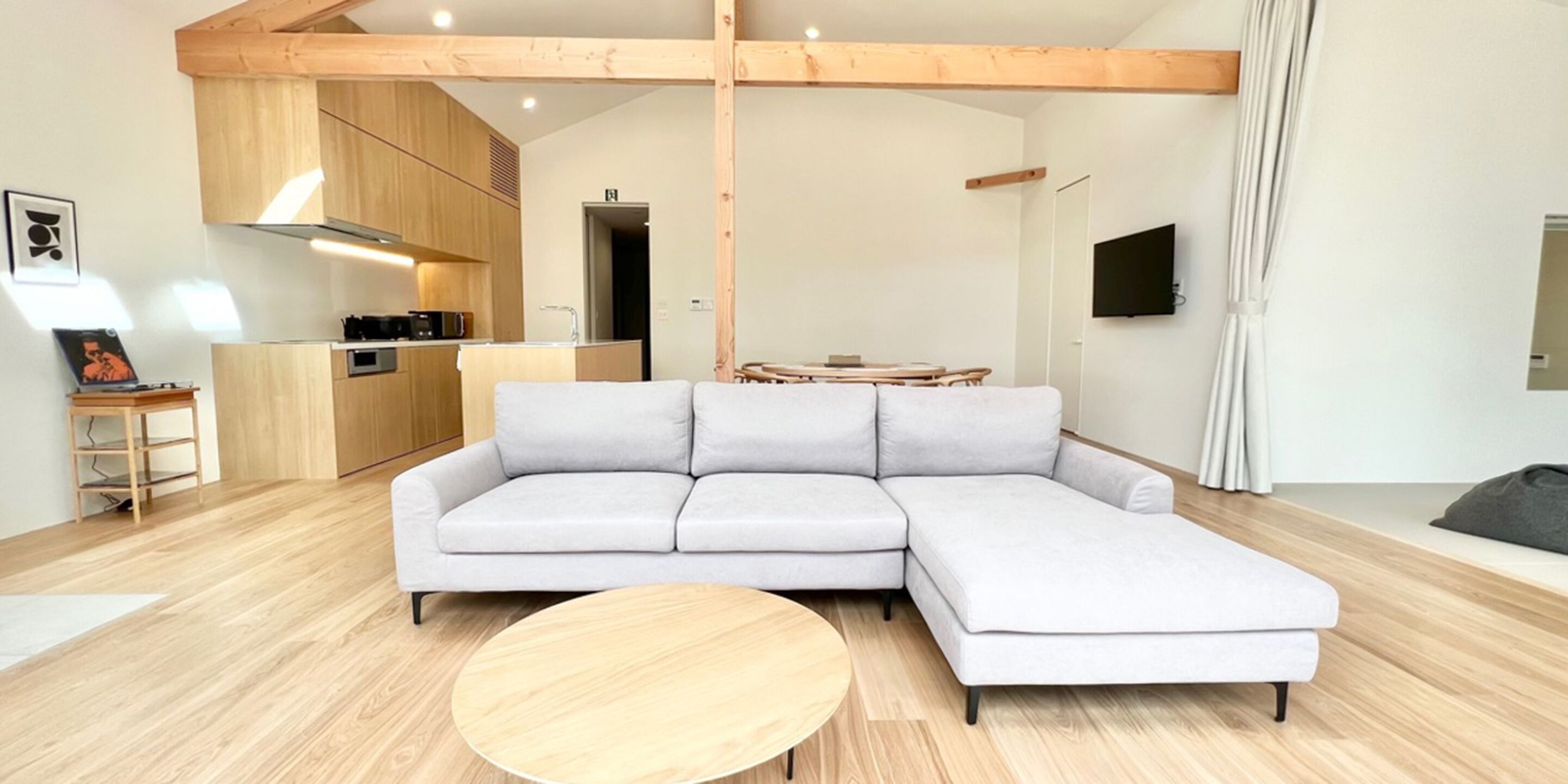 A modern living room with a light grey sectional sofa, a round wooden coffee table, and a kitchen area in the background.