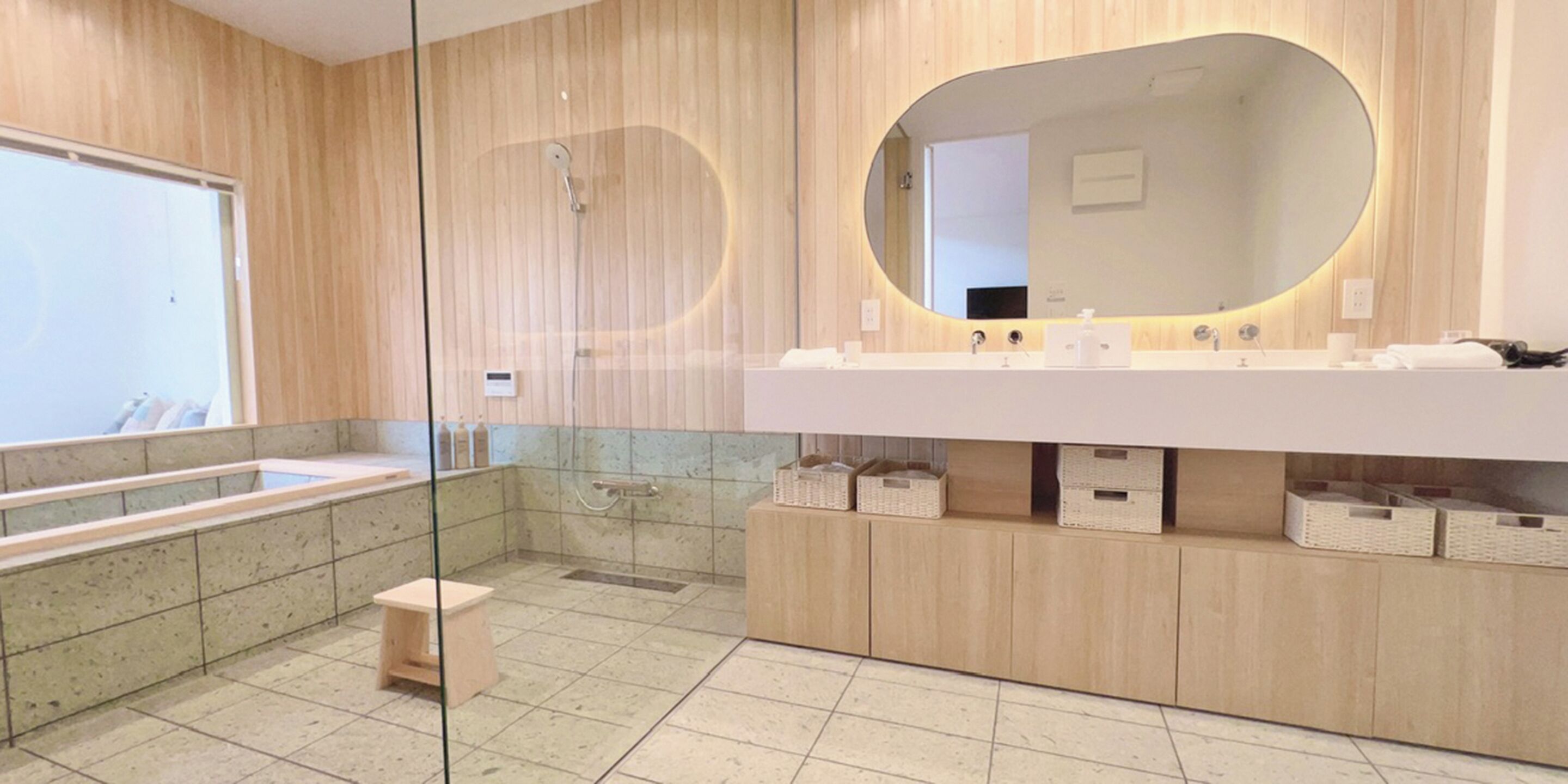 A modern bathroom with a large mirror, a bathtub, and a shower. There are wicker baskets under the sink and a small stool.