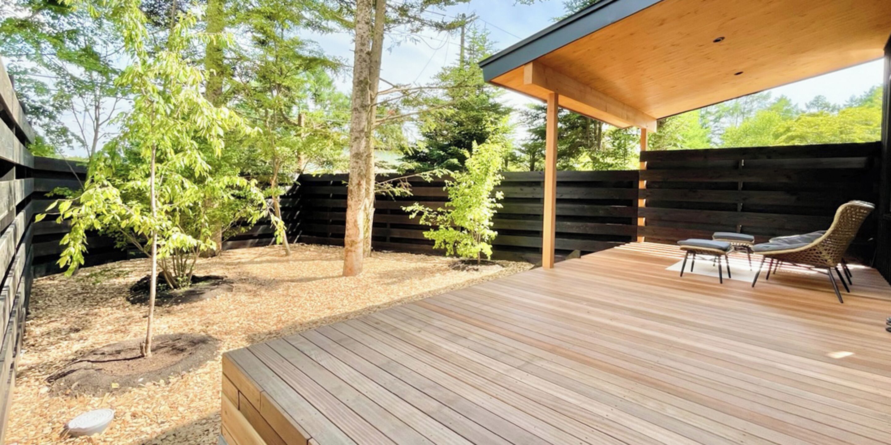 Une terrasse en bois dotée d’un espace salon abrité, entourée d’arbres et d’une clôture en bois.