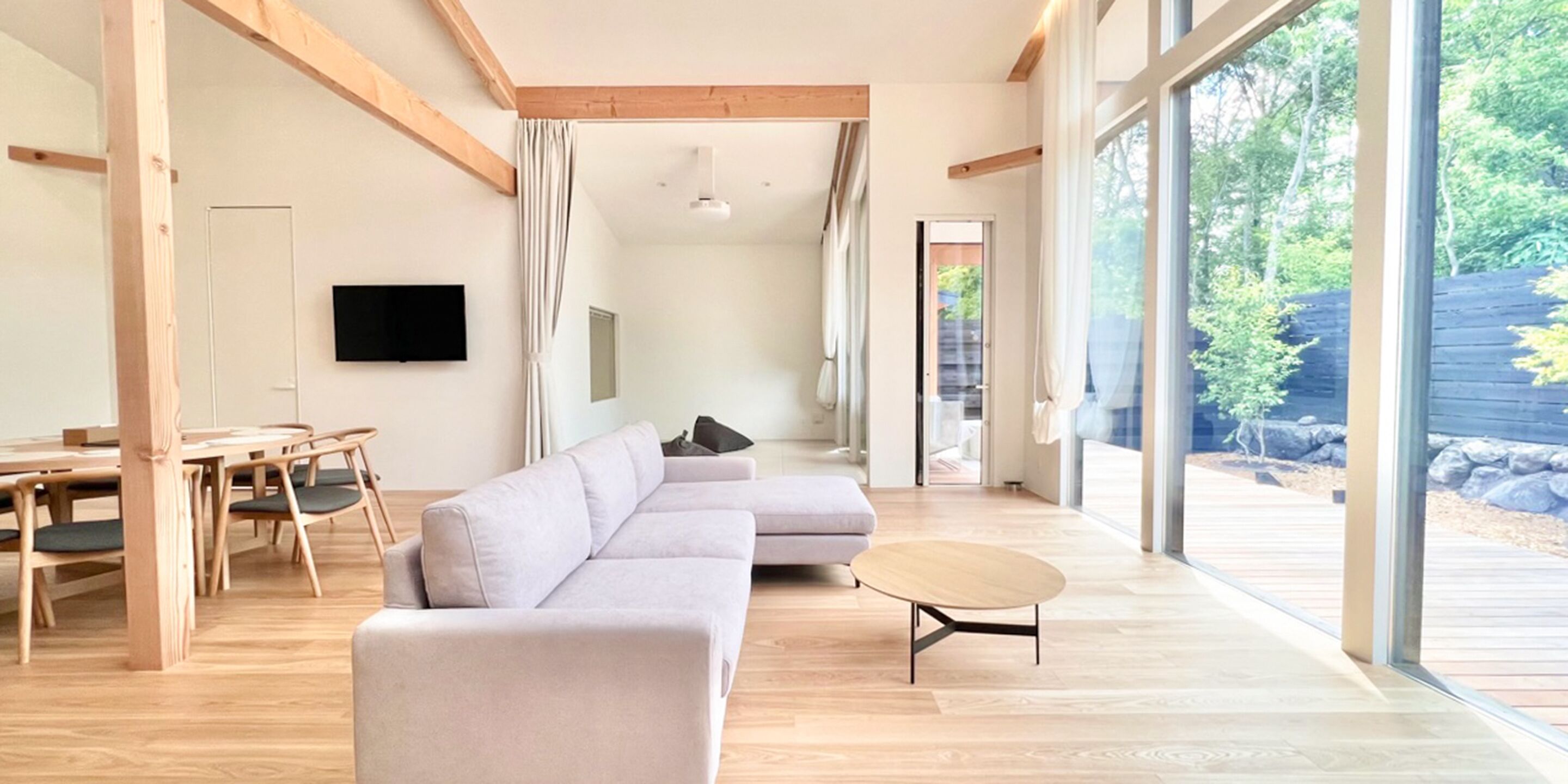 A spacious living area with a large L-shaped sofa, a round coffee table, and a dining table with chairs. There is a flat-screen TV mounted on the wall and a large window providing a view of the outdoors.