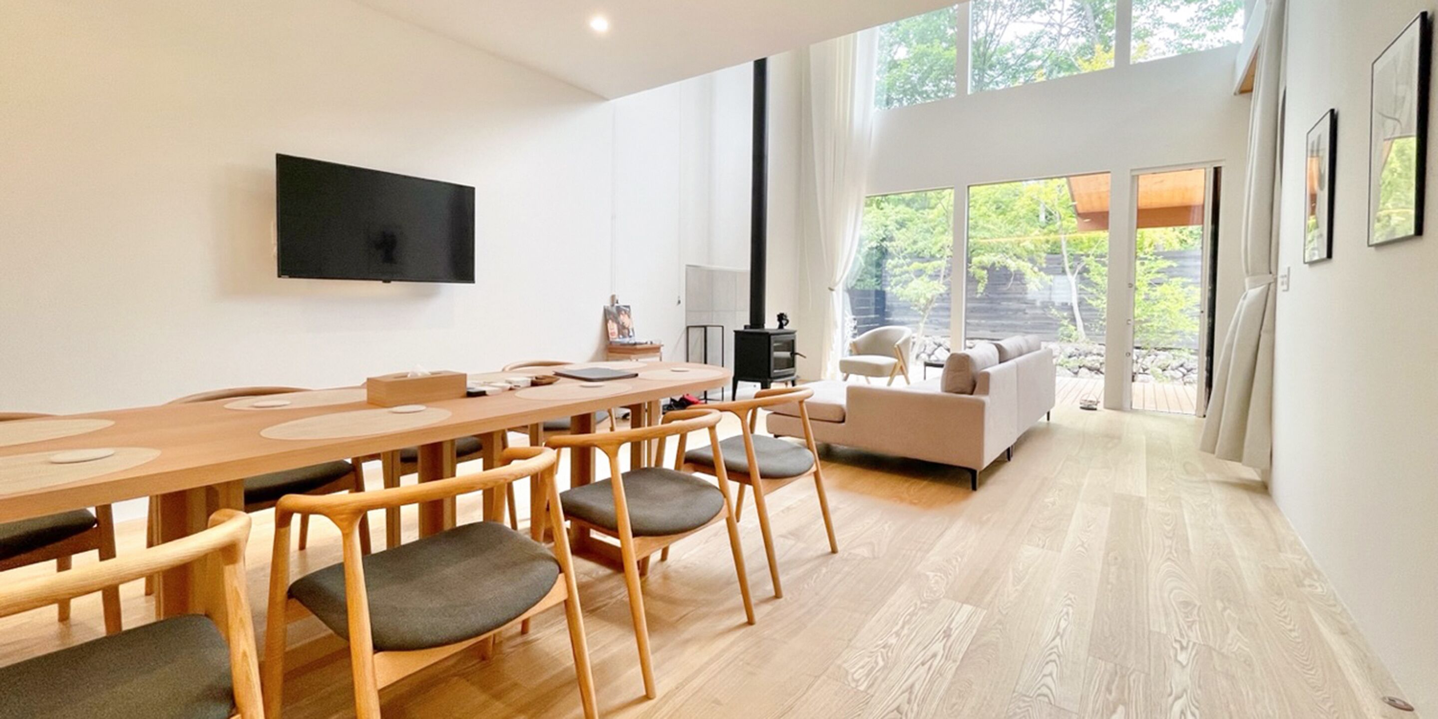 A modern living room with a wooden dining table, chairs, a sofa, and a TV mounted on the wall.