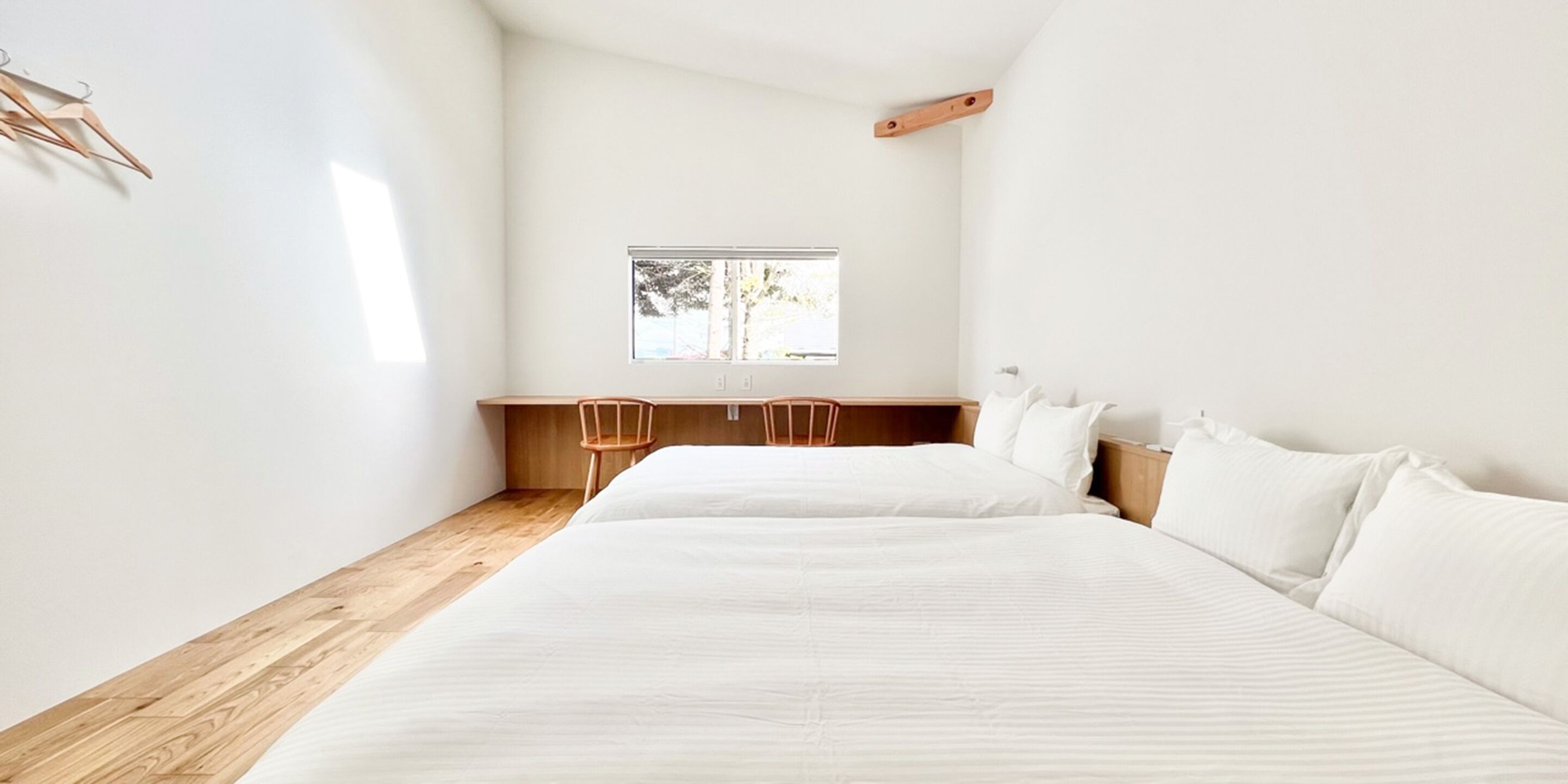 A minimalist hotel room with two beds, a wooden desk, and a window with a view of trees.