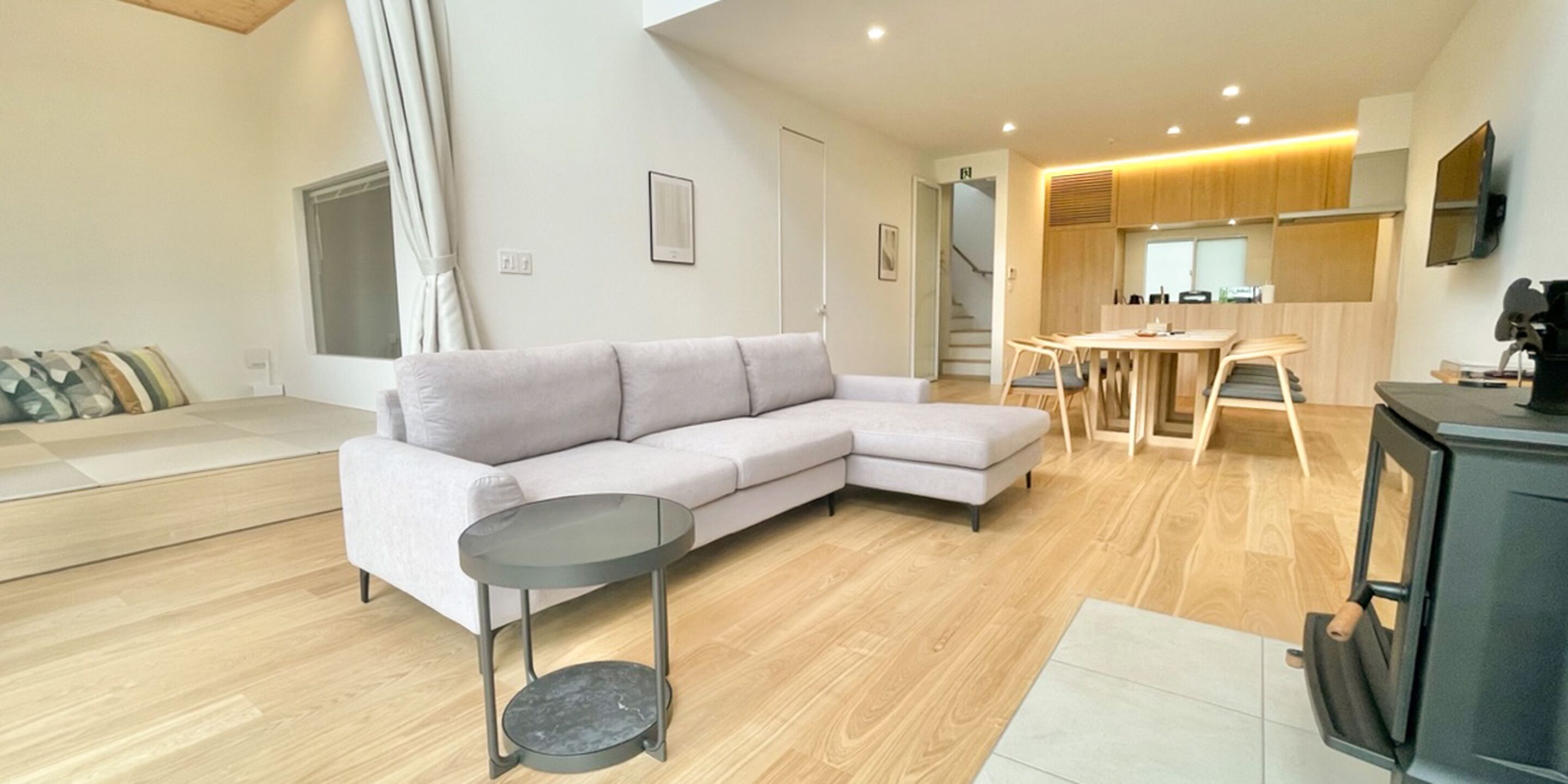 A modern living room with a grey sectional sofa, a small round table, and a kitchen area with wooden cabinets and a TV.
