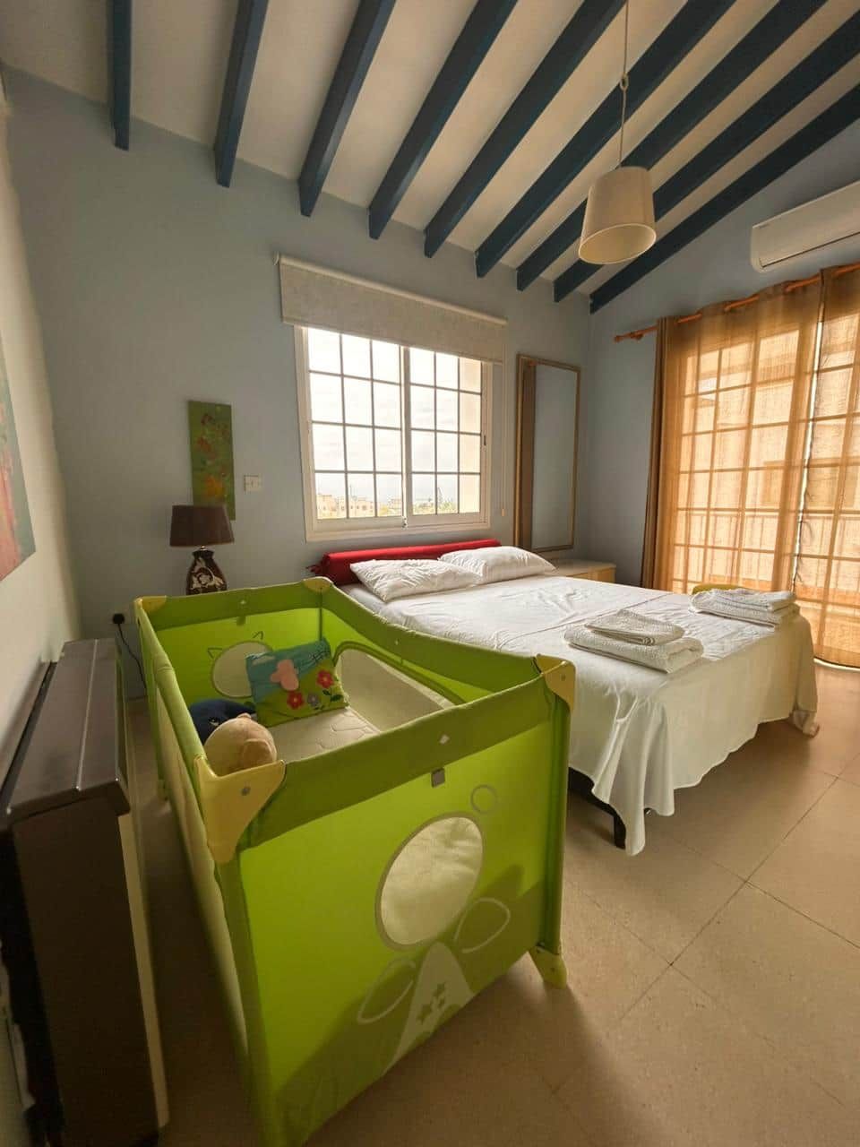 Ein Zimmer mit einem Bett, einem Kinderbett, einem Fenster und einer Decke mit freiliegenden Balken.