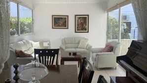 Ett vardagsrum med ett piano, två fåtöljer, ett matbord med en glasskiva och två inramade tavlor på väggen.