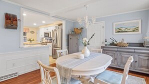 Zona pranzo con tavolo bianco, sedie e lampadario. Si nota una cucina sullo sfondo con armadietti ed elettrodomestici.