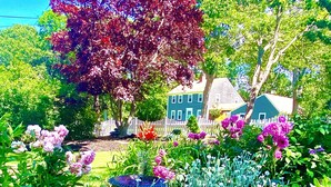 Un giardino con diverse piante e fiori, una recinzione bianca in legno a pali e una casa sullo sfondo.