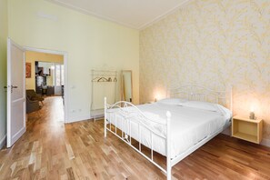 Un dormitorio con estructura de cama metálica blanca, suelo de madera, una mesita de noche con lámpara y papel tapiz floral.