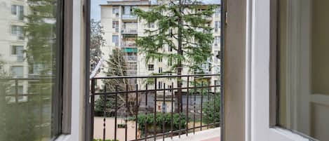 A balcony with a view of a multi-story building, a metal fence, and some greenery.