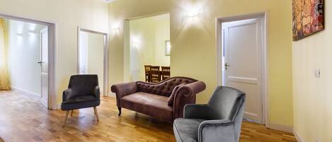 A spacious living room with hardwood flooring, a brown leather sofa, a grey armchair, and a wooden dining table with chairs.