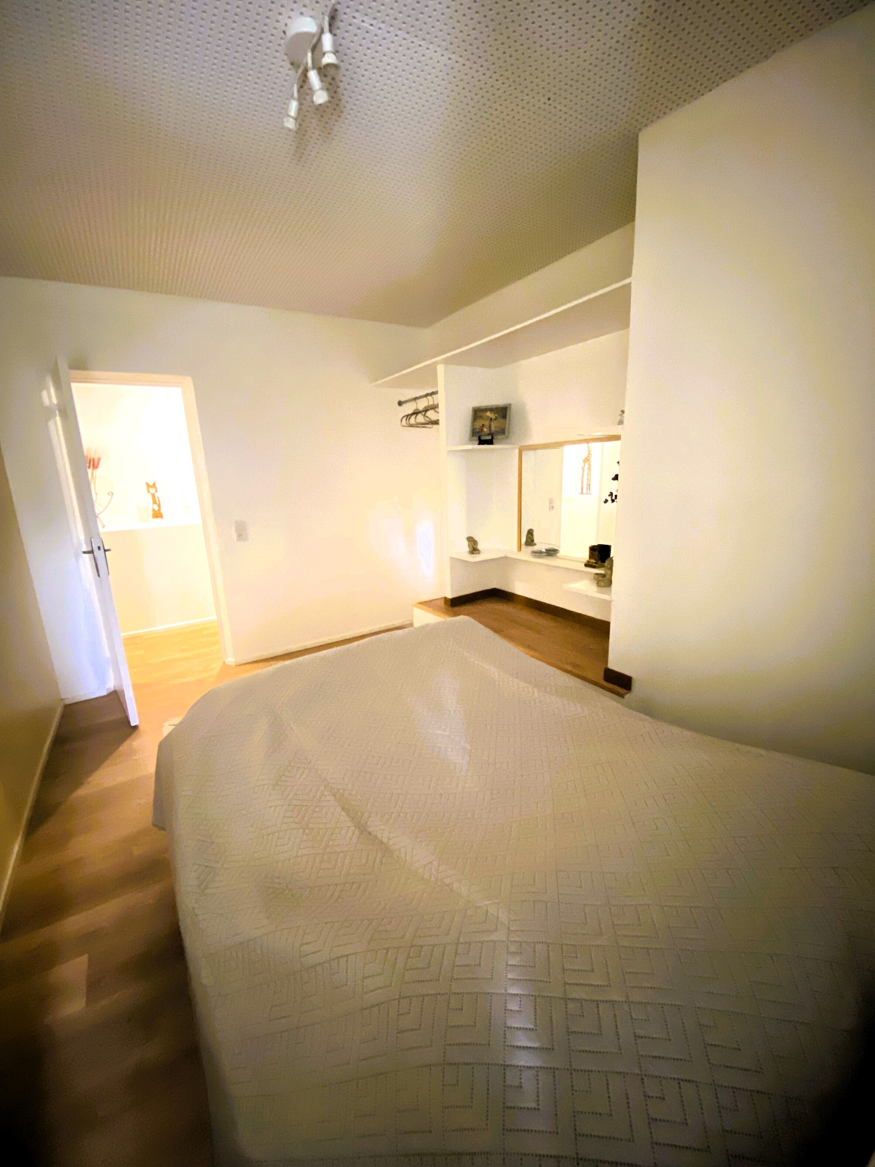 Een slaapkamer met een groot bed, een houten vloer, een wit plafond en een deur naar een andere kamer.