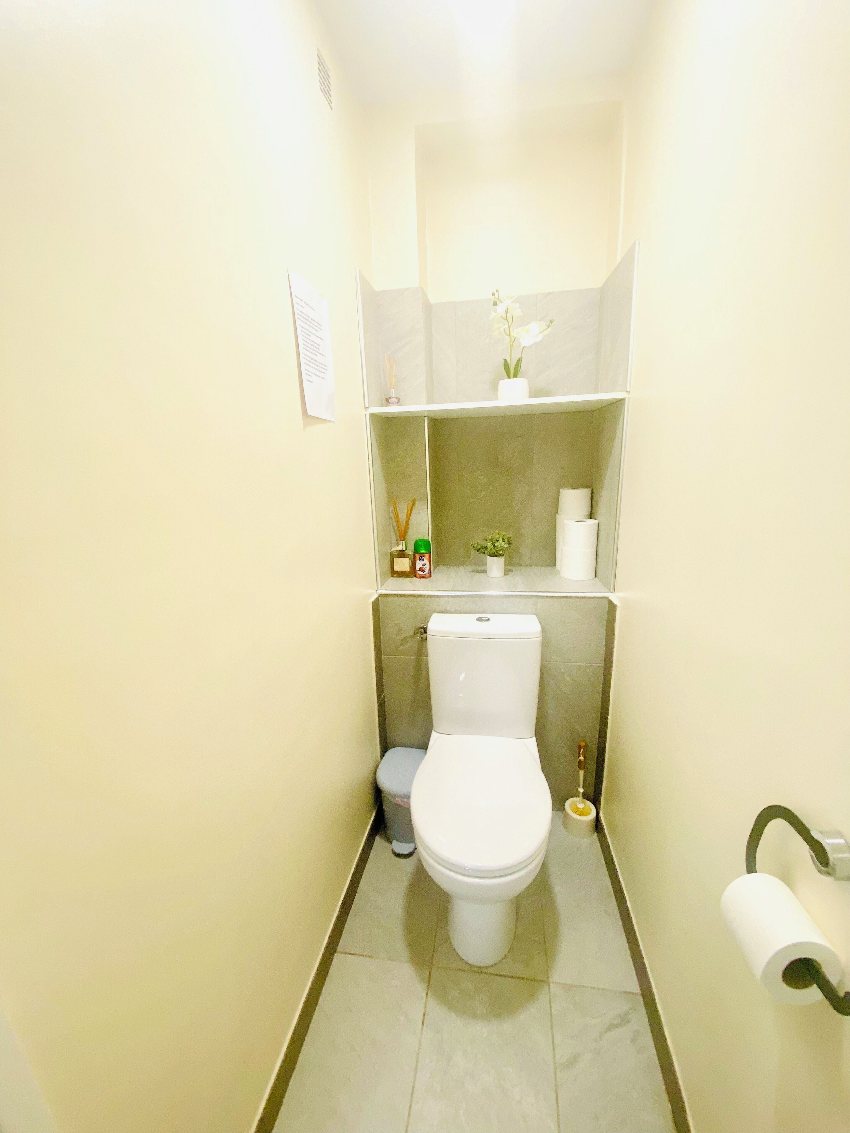 A small, narrow bathroom with a toilet, a trash bin, and a shelf above the toilet holding a small plant and toiletries.