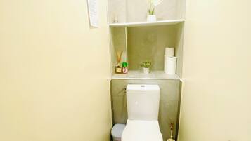 A small, narrow bathroom with a toilet, a trash bin, and a shelf above the toilet holding a small plant and toiletries.