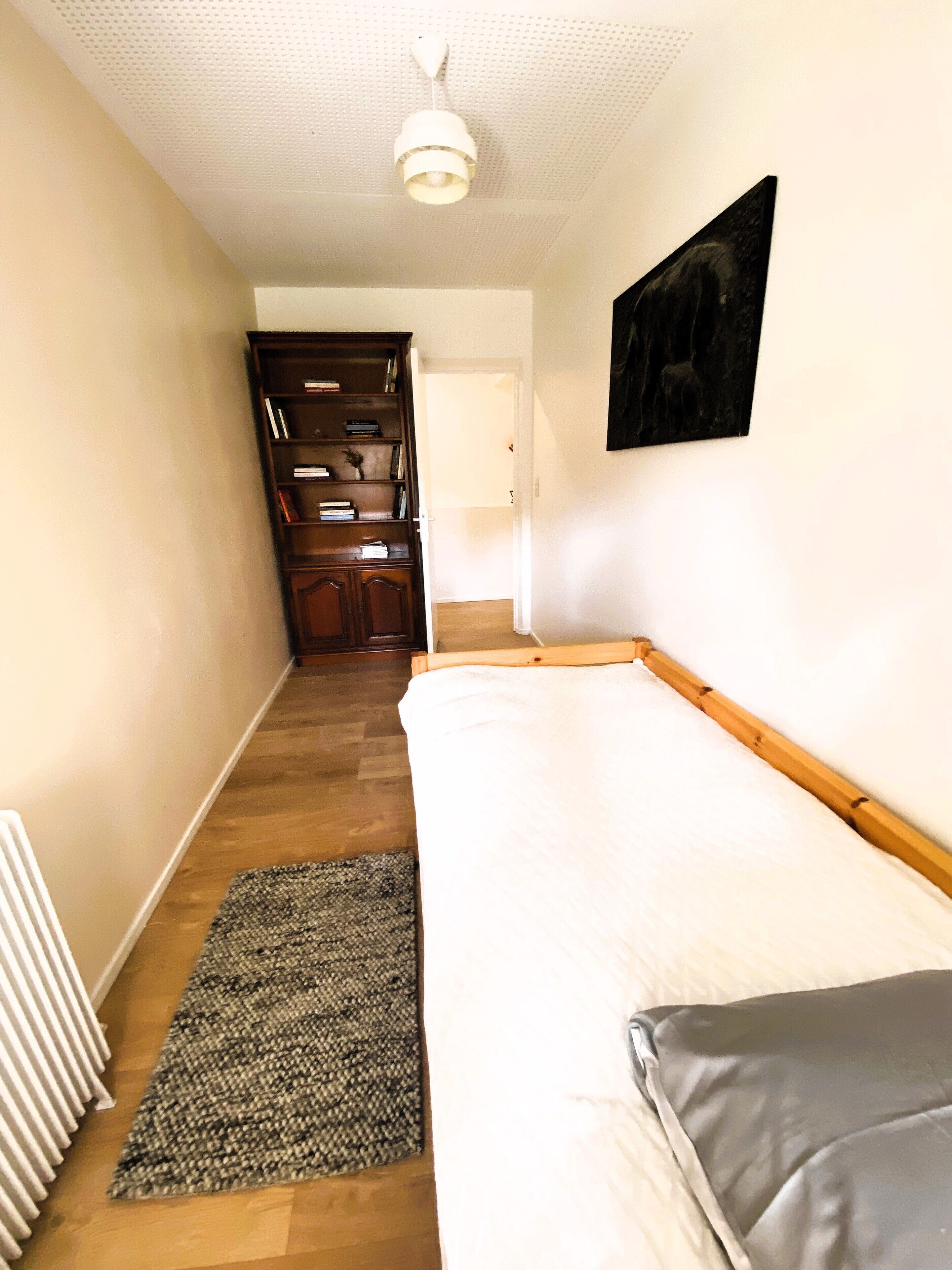 Una habitación estrecha con una cama, un radiador, suelo de madera, una estantería y una puerta que da a otra habitación.