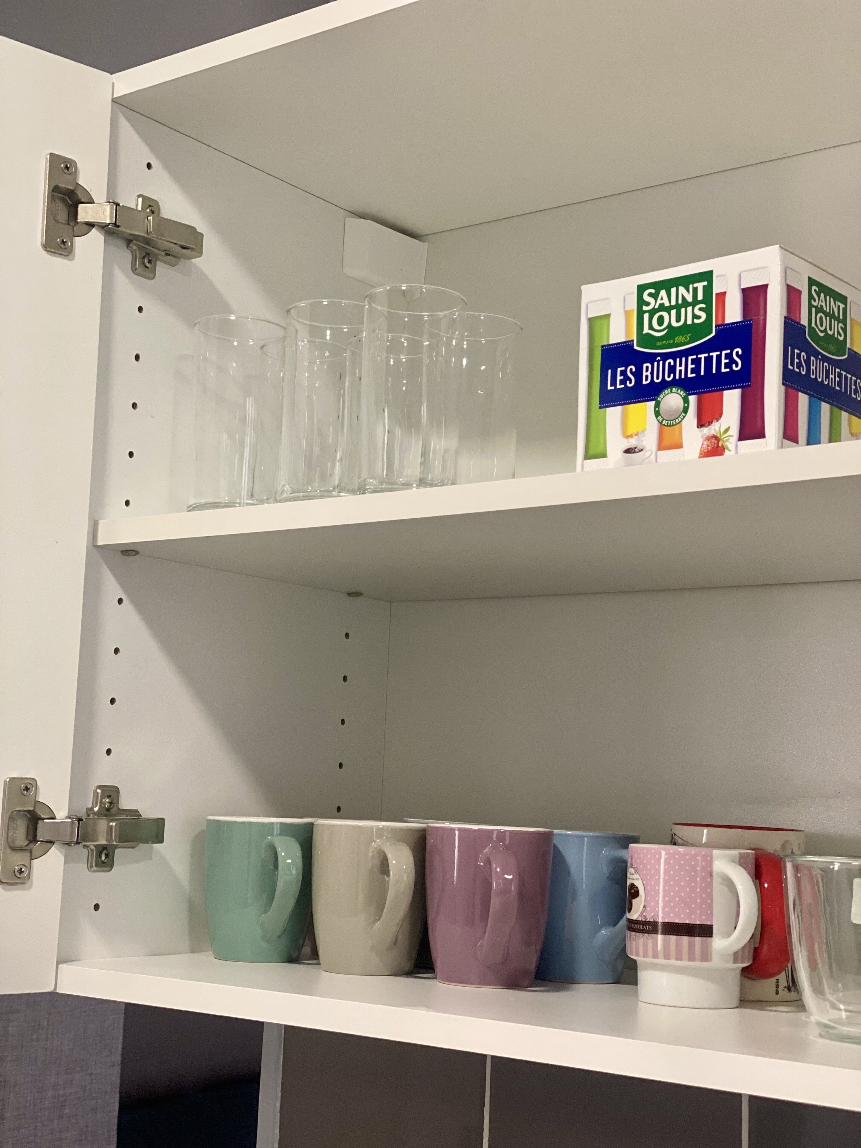 A kitchen cabinet with clear drinking glasses on the upper shelf and colorful ceramic mugs on the lower shelf.