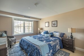 A bedroom with a large bed, two bedside tables, a chair, a window, and a ceiling vent. - Condo Room 300 Grand Victorian's Largest Rental! (Old Orchard Beach)