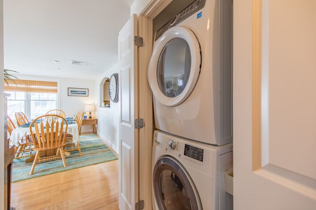 A laundry room with stacked washers and dryers, a dining area with wooden chairs and a table, and a window with blinds.