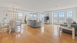 A spacious living room with a grey sofa, a dining table with chairs, a TV, and a large window with a view of the sea. - Direct Ocean Front Unit With Private Balcony. (Old Orchard Beach)