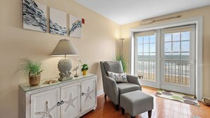 Une pièce avec un fauteuil gris, un meuble blanc orné de motifs d’étoiles de mer, une lampe et une vue sur la plage, visible à travers une porte coulissante.