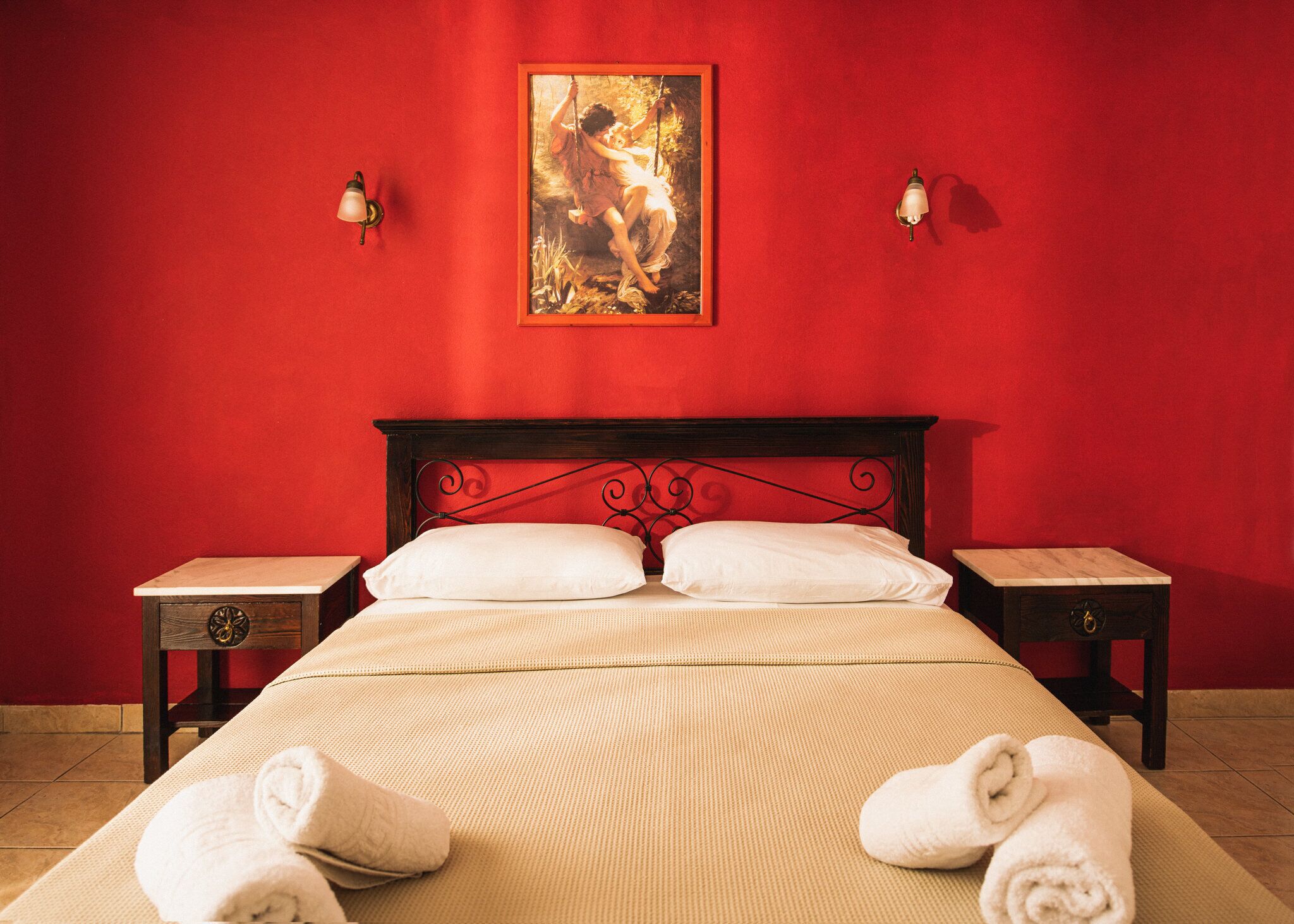 A hotel room with a red wall, a bed with white linens, two bedside tables, and a painting above the bed.