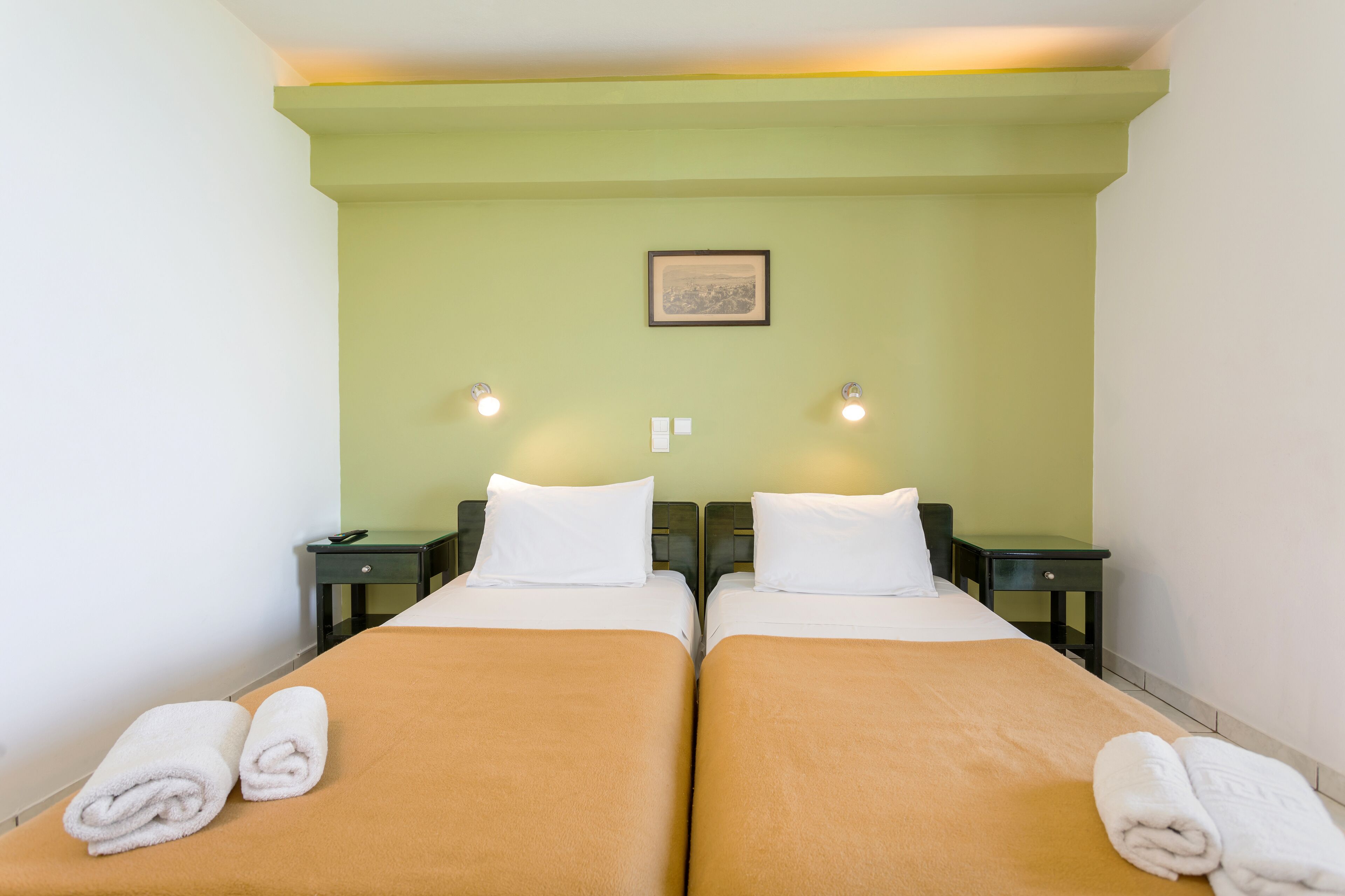 A hotel room with two beds, each with a brown bedspread and white pillows. There are two bedside tables with lamps, a framed picture on the wall, and a small nightstand with a phone.