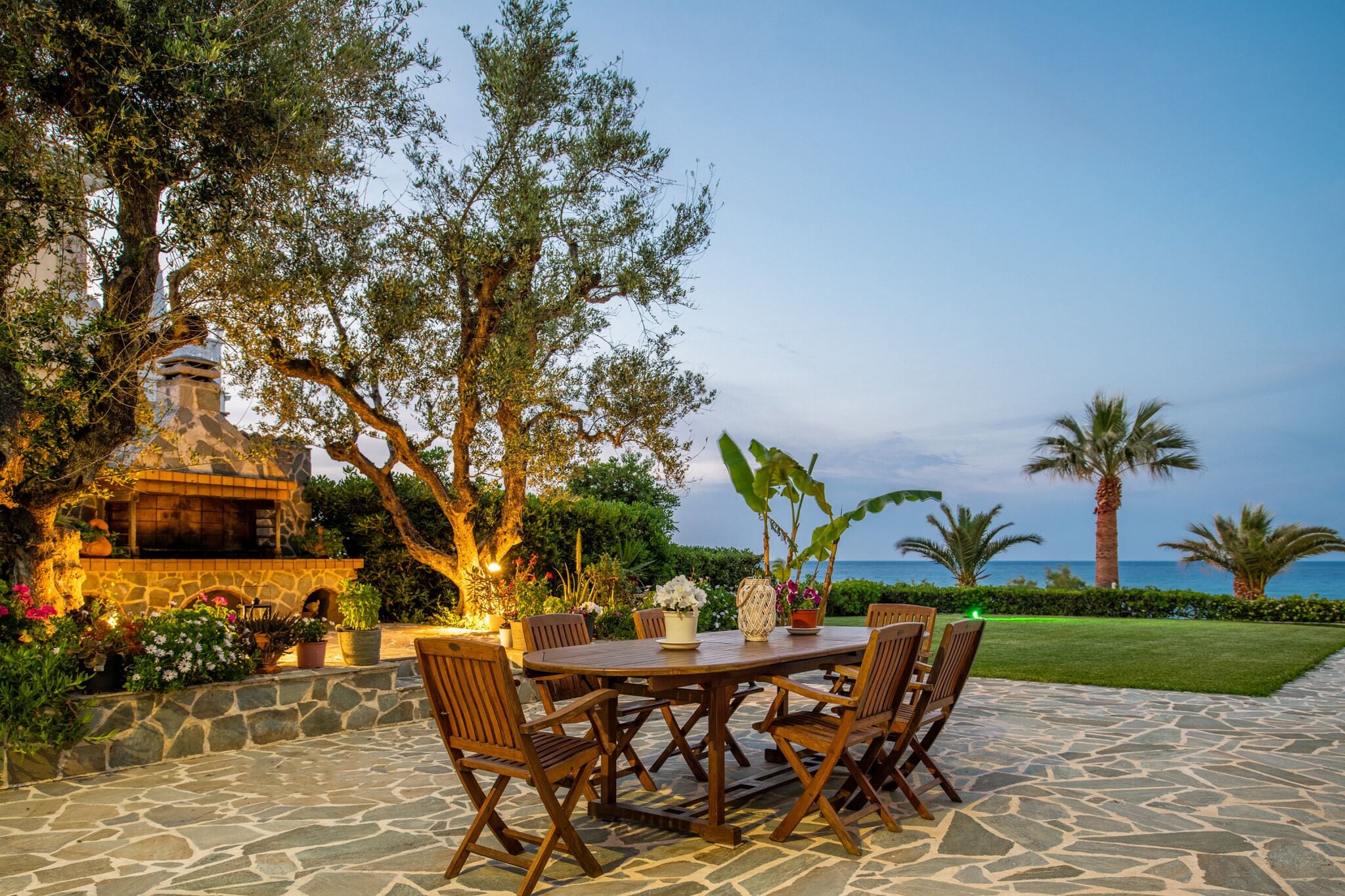 An outdoor dining area with a stone table and wooden chairs, surrounded by lush greenery and a view of the sea.