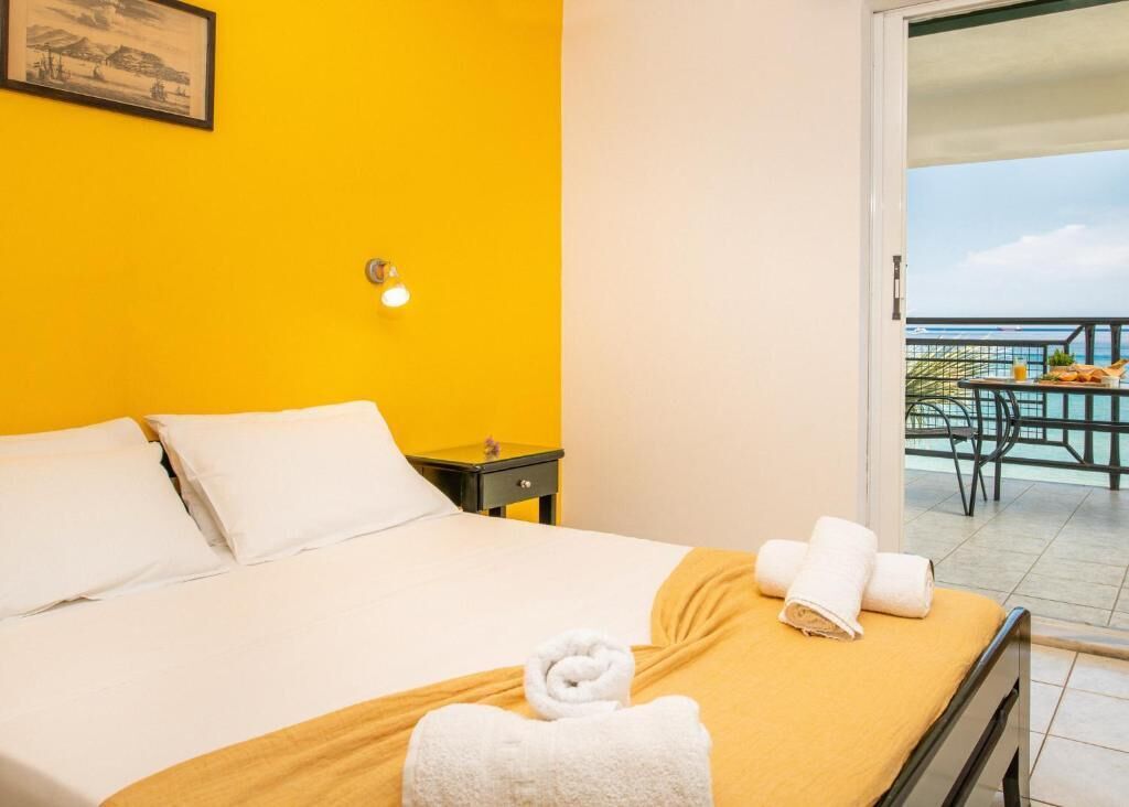 Une chambre d’hôtel avec un lit, une table de chevet, un mur jaune, un tableau encadré et un balcon avec une table et des chaises.
