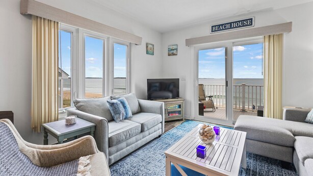 Un salón amplio con un televisor grande, asientos cómodos, un balcón con vistas a la playa y un letrero que dice "BEACH HOUSE".
