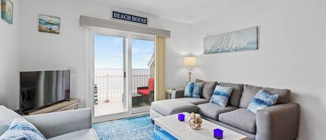 Ett vardagsrum med en soffa, en tv, ett bord och en balkong med en skylt som visar texten ”BEACH HOUSE”.