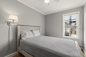 A bedroom with a bed, a nightstand, a lamp, and a window with a view of buildings. - Seaside Village Condo #3- 300 ft to Sandy Beaches! (Old Orchard Beach)