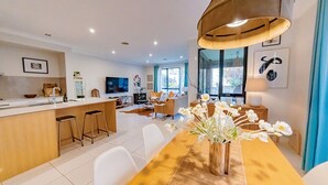 A modern kitchen with a central island, a dining area with a wooden table and white chairs, and a large window providing natural light. - StayAU 4BR Designer Home in Knox (Wantirna South)