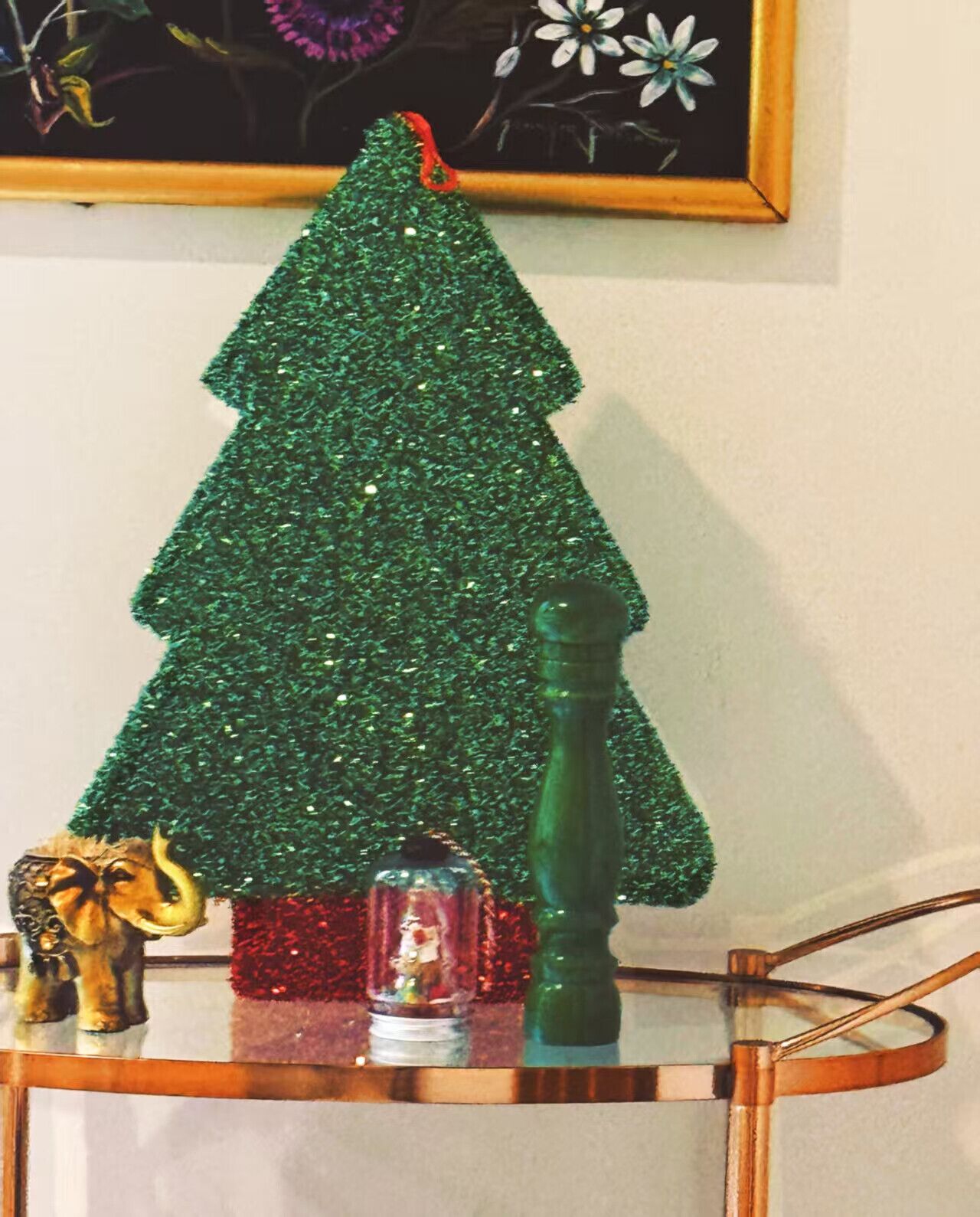 A small green Christmas tree on a glass table with a red ornament and a glass bottle.