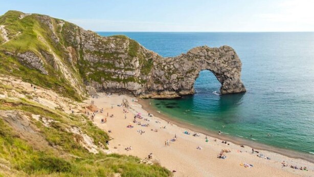 Un paisaje costero con un arco natural de roca, una playa de arena y aguas azules cristalinas.