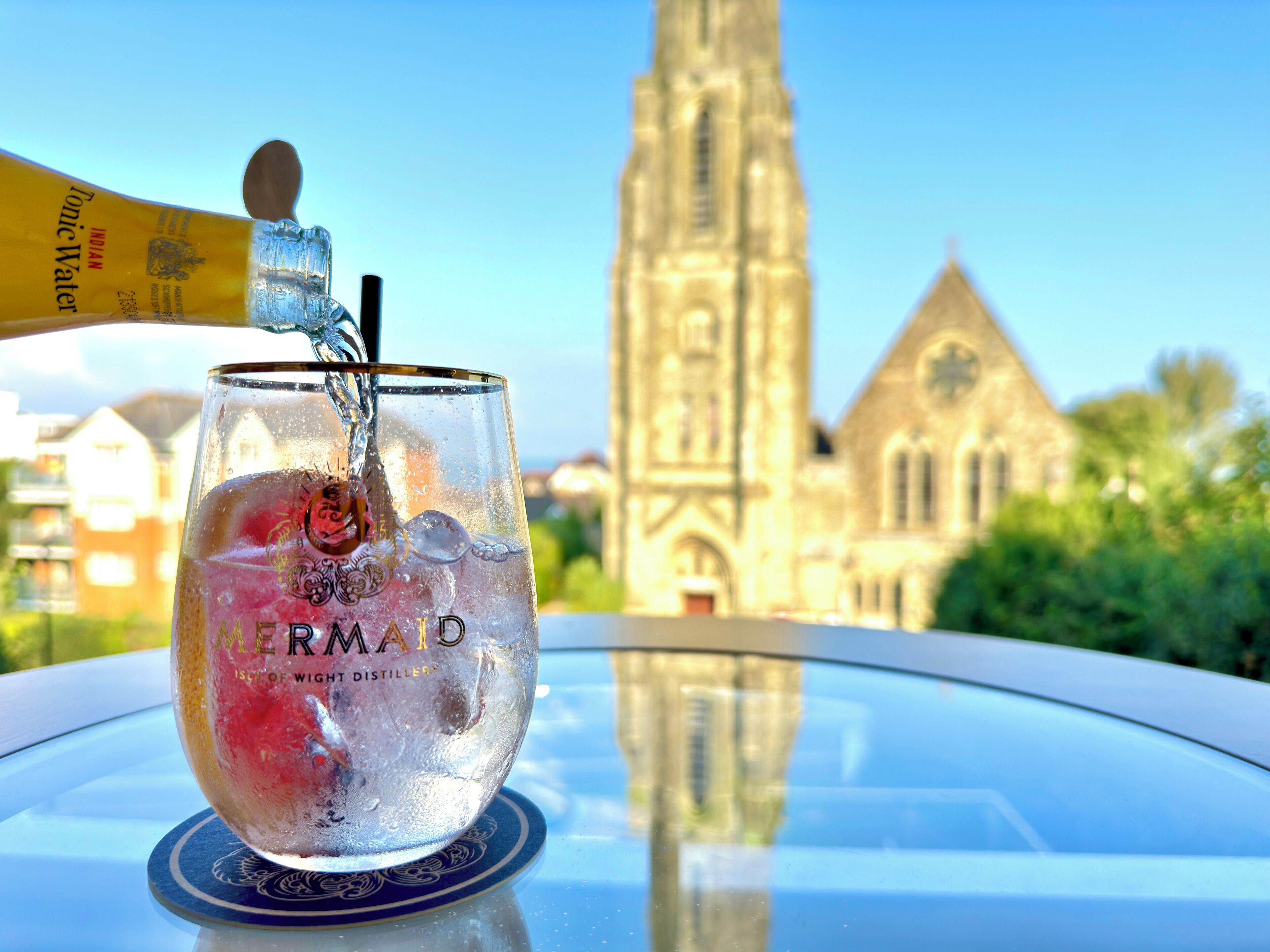 Un vaso de agua con cubitos de hielo y una botella de agua tónica sobre una mesa, con vistas a una iglesia al fondo.