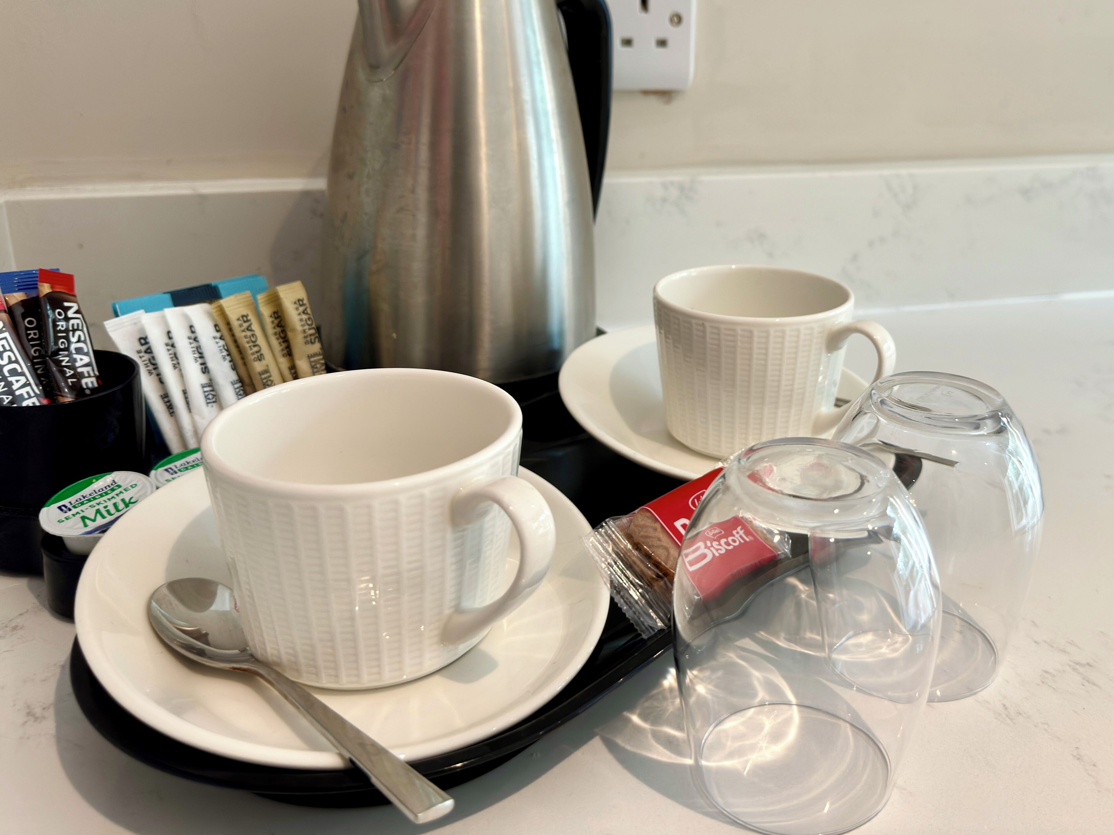 Una presentación para el desayuno con dos tazas blancas, una cuchara, un hervidor de té y una cafetera sobre una encimera.