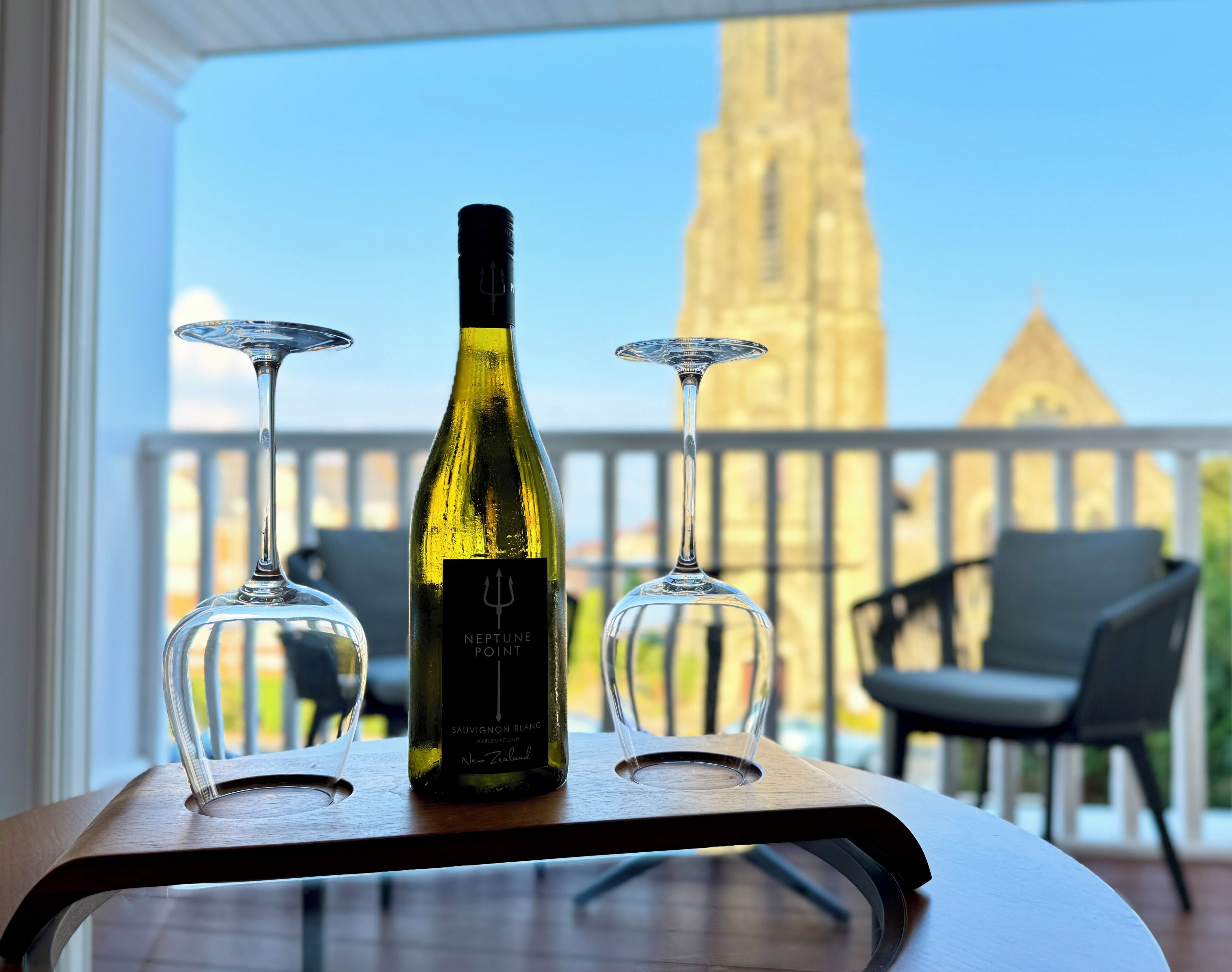 A bottle of wine and three glasses on a table with a view of a tower and a building in the background.