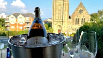 A bottle of JULES FERAULT champagne in a metal bucket with ice cubes, two empty wine glasses, and a view of a church and residential buildings in the background.