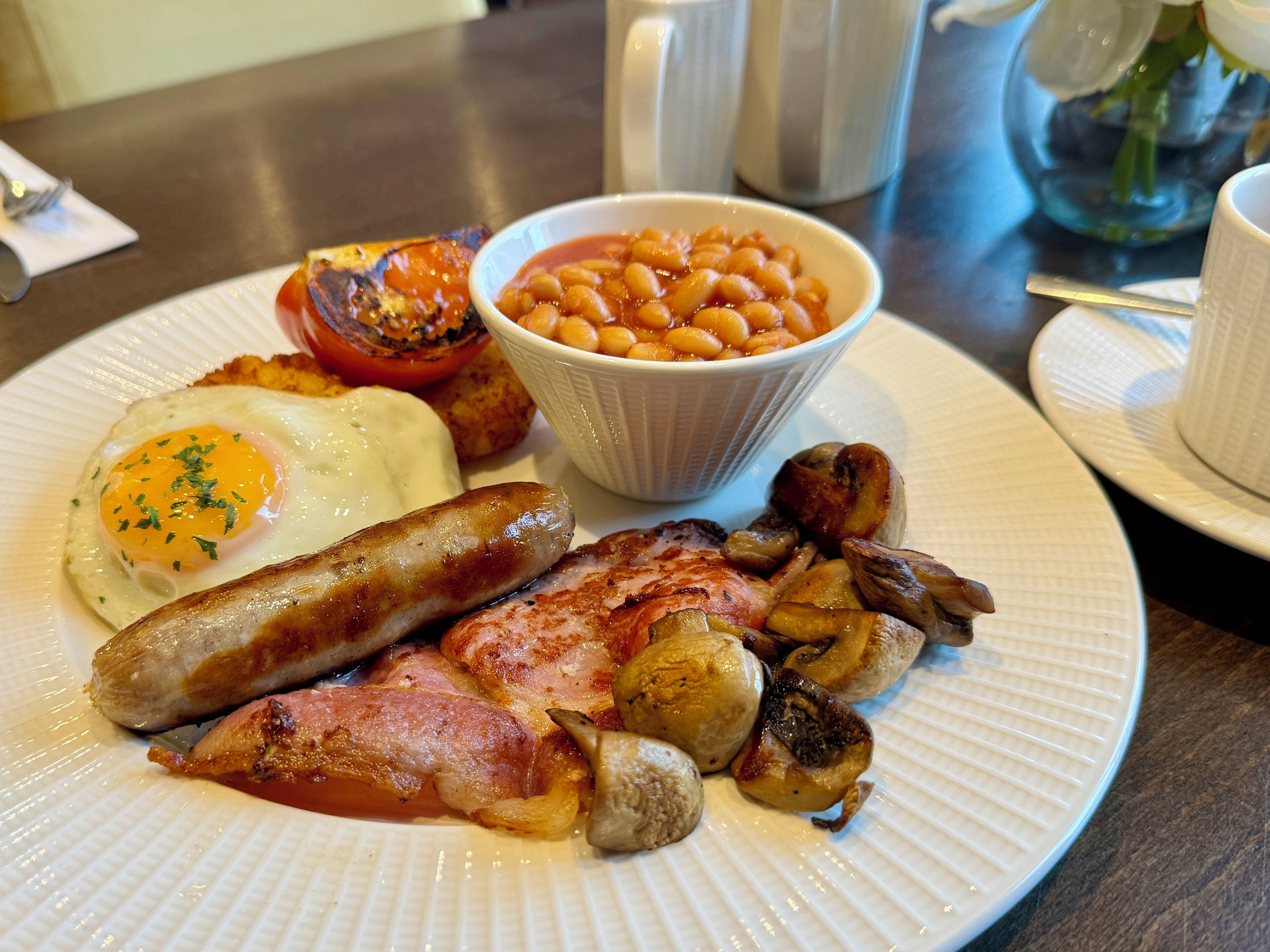 A plate of breakfast food including eggs, sausages, bacon, mushrooms, and baked beans.