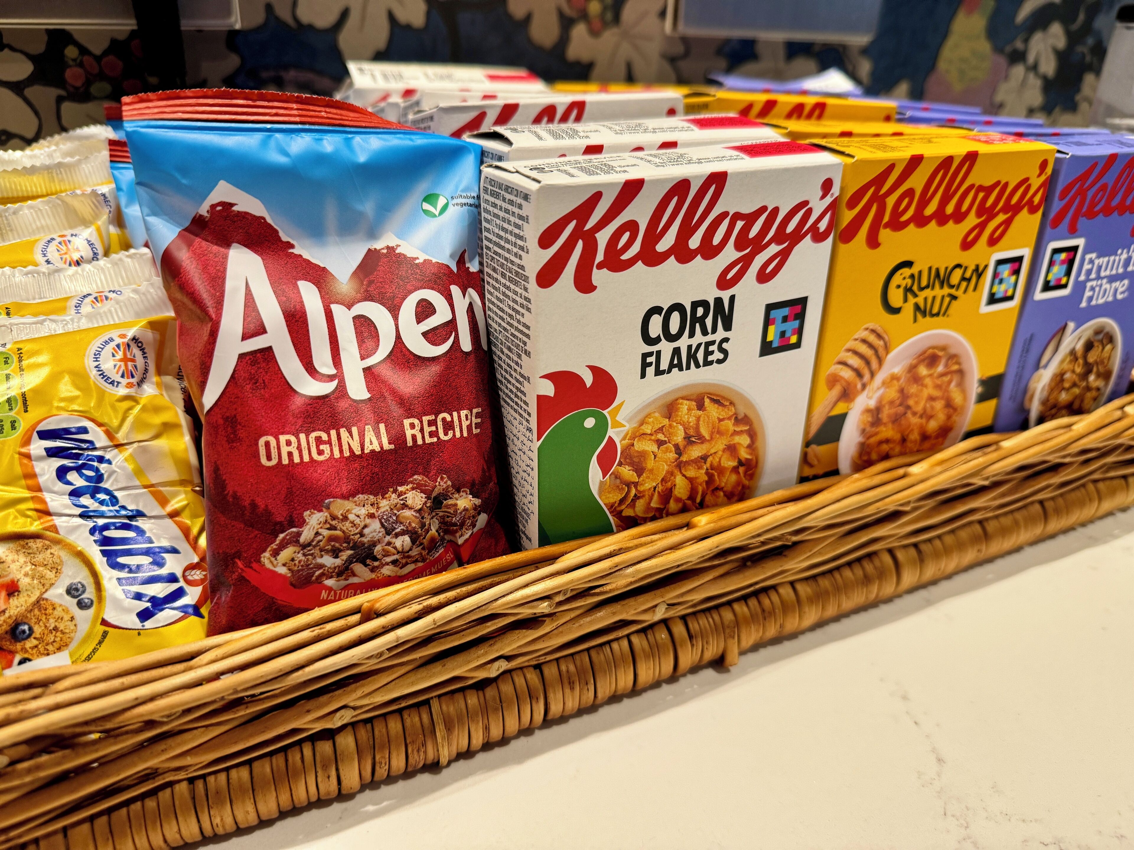 A variety of cereal boxes including Alpen, Kellogg's Corn Flakes, and Fruit'n Nut, arranged on a wicker tray.