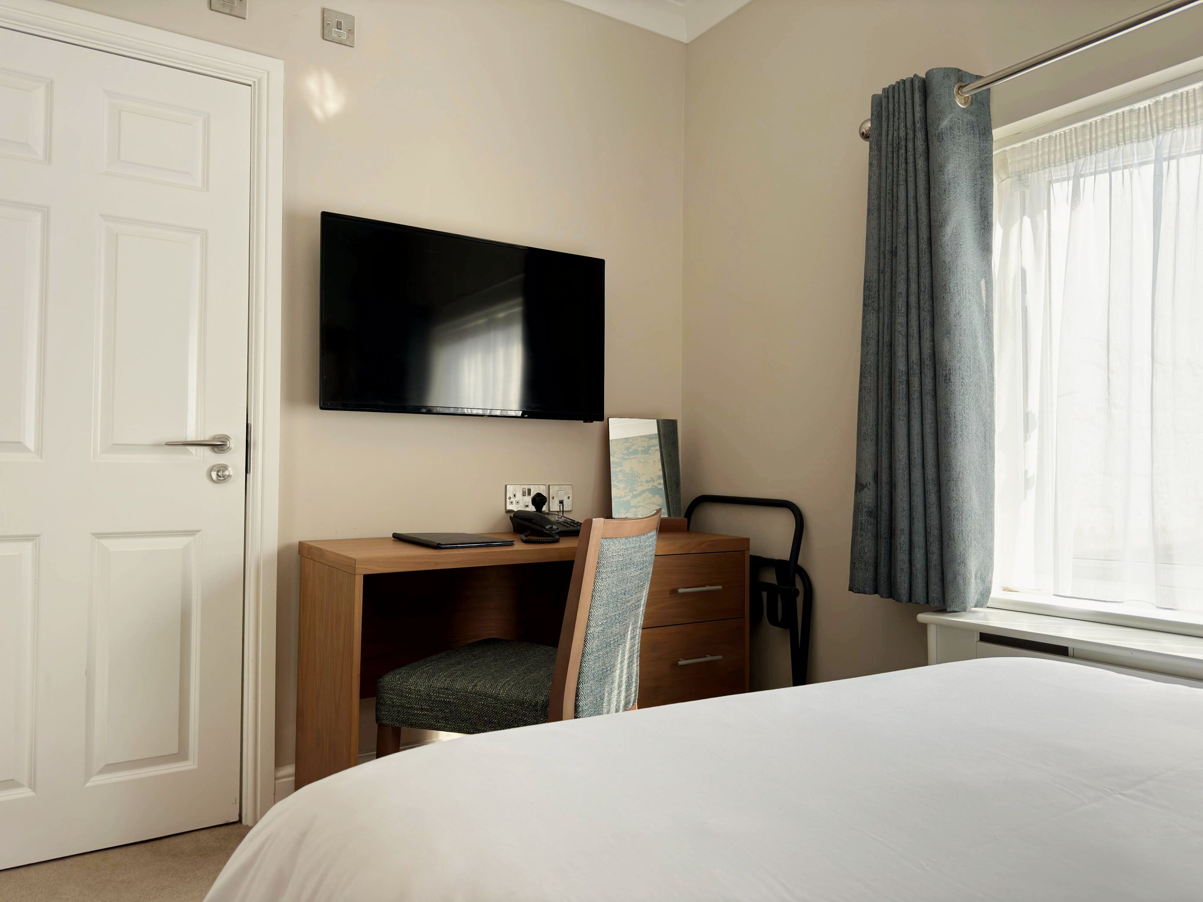 A hotel room with a bed, a desk with a chair, a television, a wardrobe, and a window with curtains.