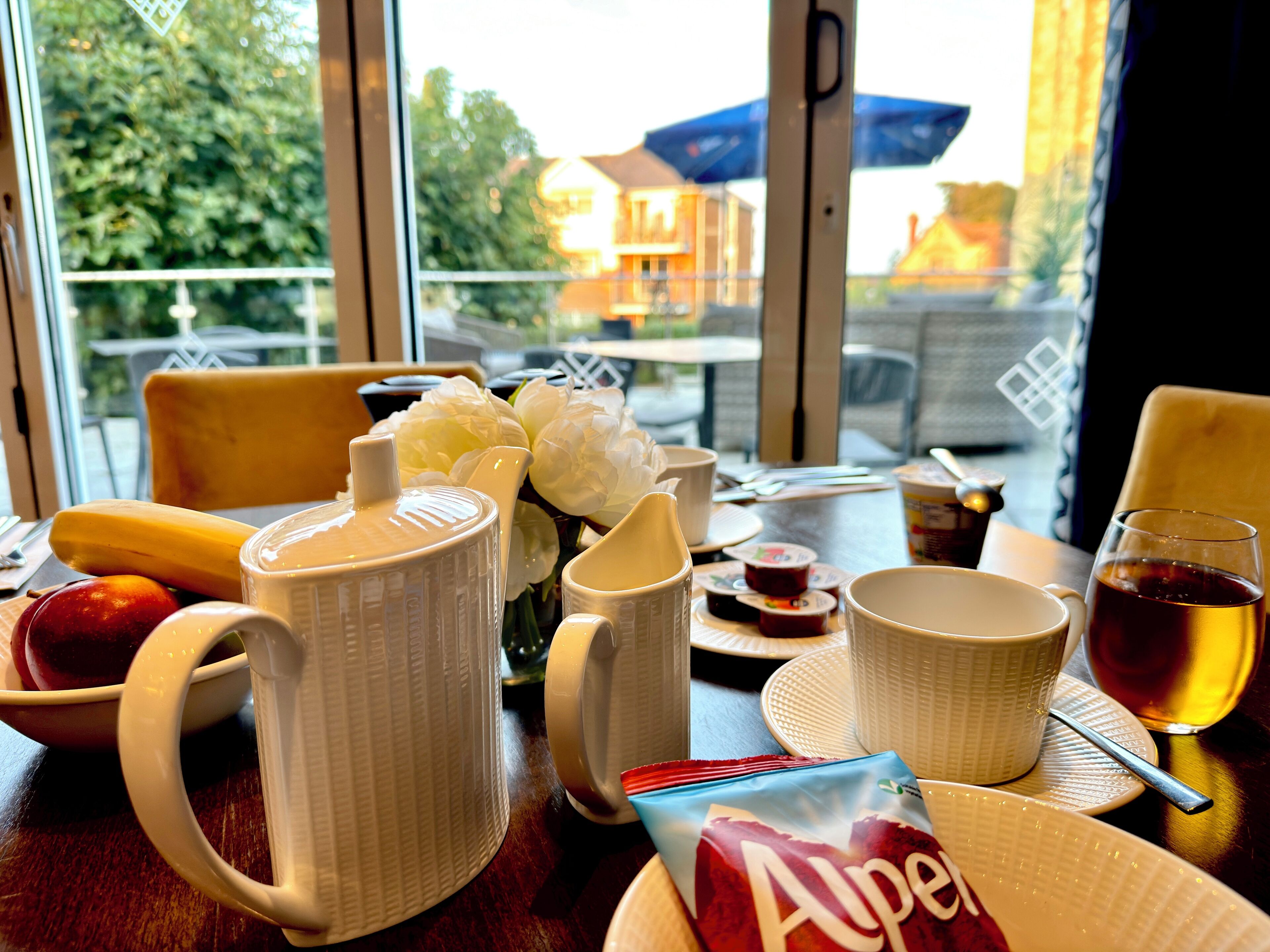 A table with breakfast items including a teapot, cups, a plate with a bag of chips, and a glass of tea.
