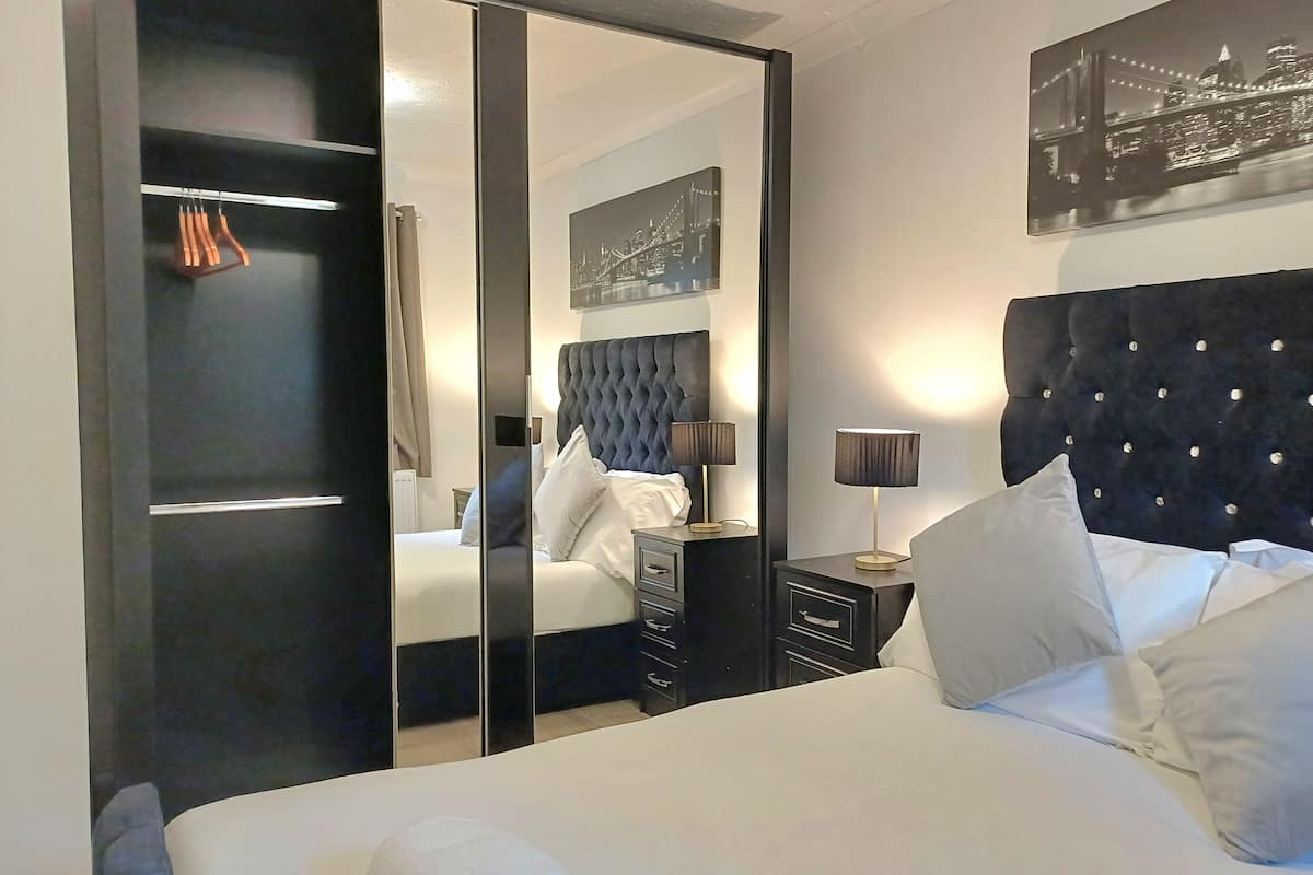 A hotel room with a large bed, a wardrobe, a bedside table with a lamp, and a cityscape painting on the wall.