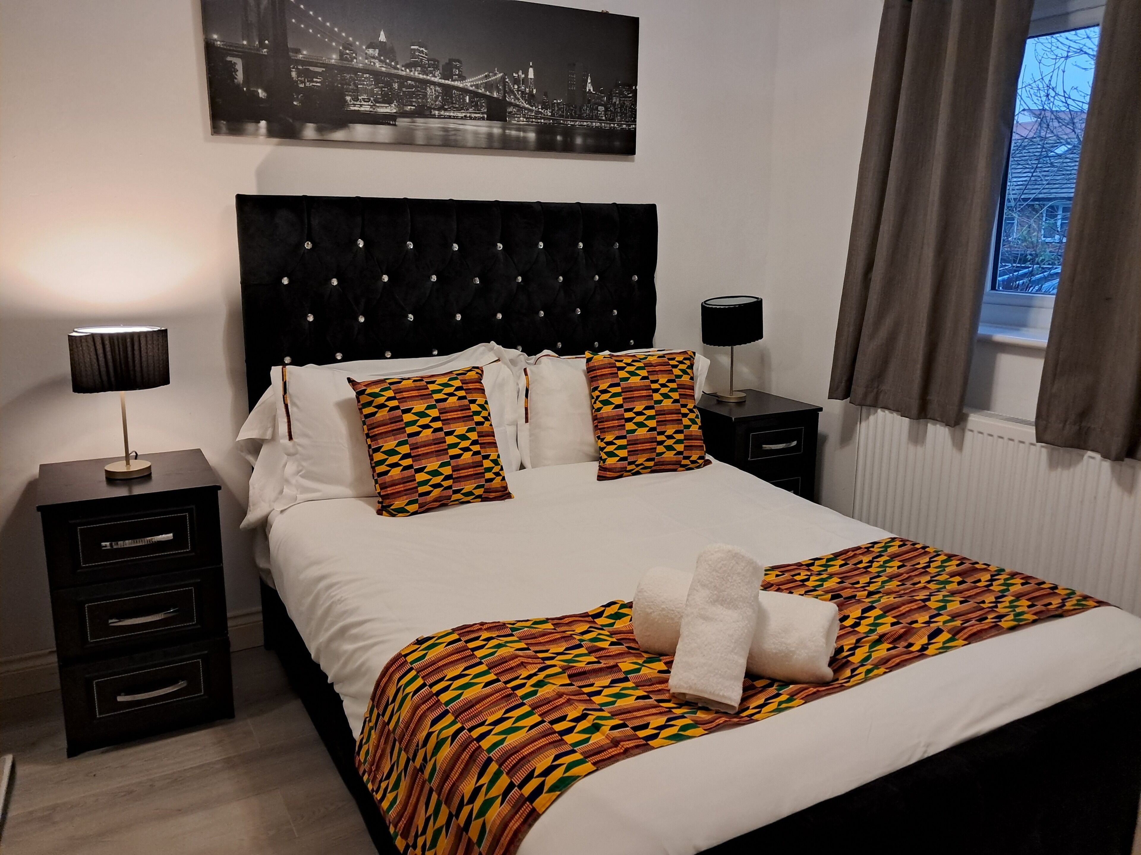 A neatly made bed with patterned pillows, a black headboard, and a cityscape painting above.