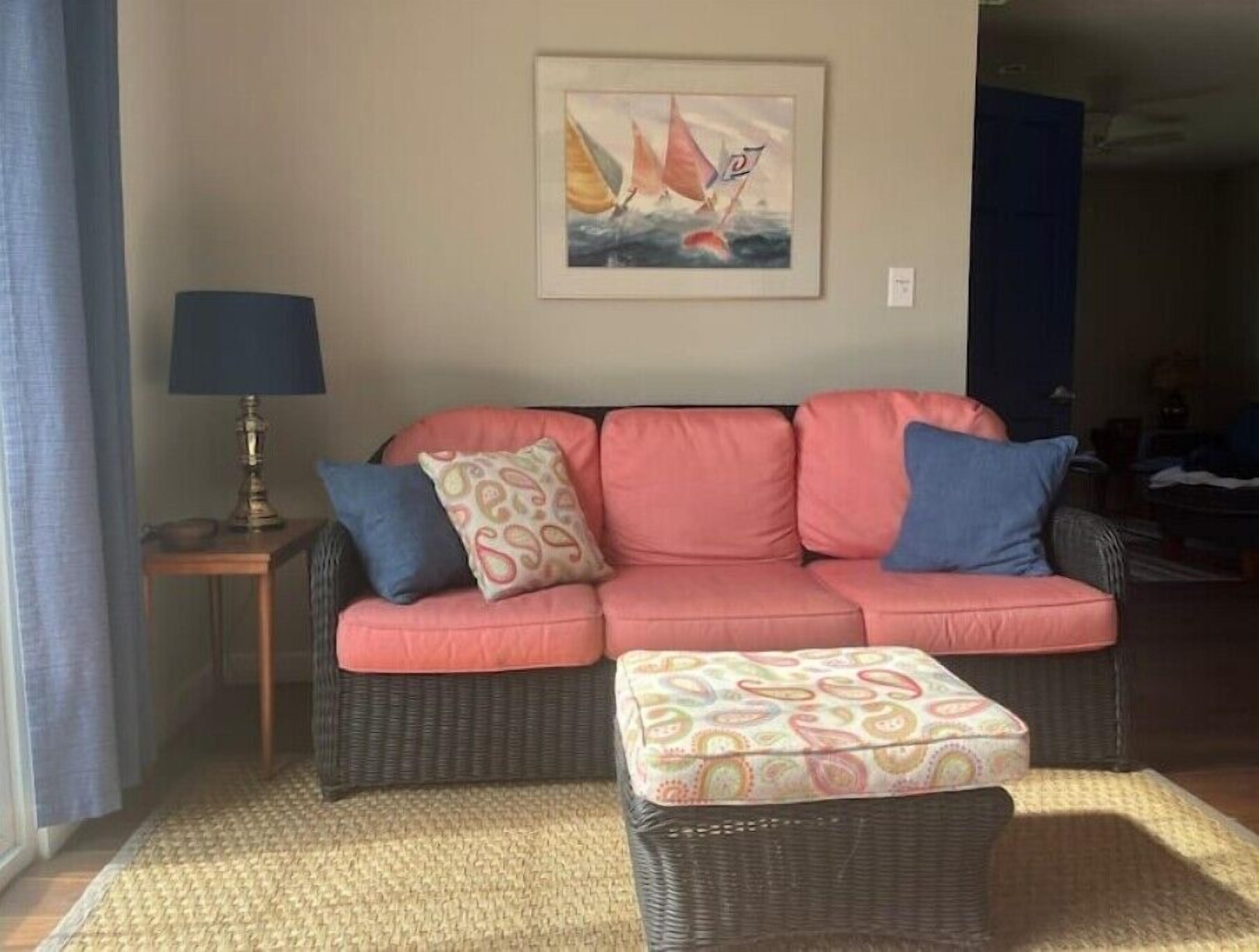 Uma sala de estar com um sofá rosa, duas almofadas estampadas, uma mesa pequena com um abajur e um quadro com barcos à vela na parede.